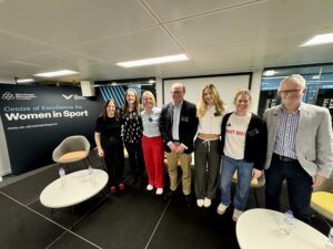 A group of people pose for the camera in front of branding that reads Centre of Excellence for Women in Sport